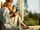 Mulher sorrindo enquanto usa o celular em um ambiente tranquilo, simbolizando o bem-estar diário com o celular.